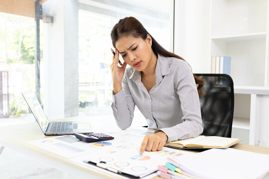 A woman is working on a desk in front of her with a pile of documents, a laptop, a calculator, a mobile phone and a notebook. This image shows her working on a project or analyzing data.
