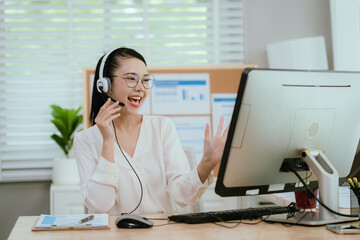 Smiling Asian female call center agent wearing a headset, speaking with a client at her office...