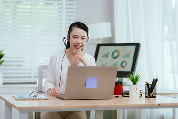 Smiling Asian female call center agent wearing a headset, speaking with a client at her office...