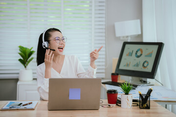 Smiling Asian female call center agent wearing a headset, speaking with a client at her office...