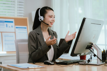 Smiling Asian female call center agent wearing a headset, speaking with a client at her office desk, providing professional customer support.