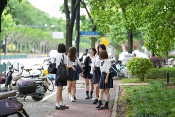 In early June in Pudong, Shanghai, seven Japanese high school girls in summer uniforms walk together down a city street, smiling and chatting as they head to school under the soft morning sunlight.