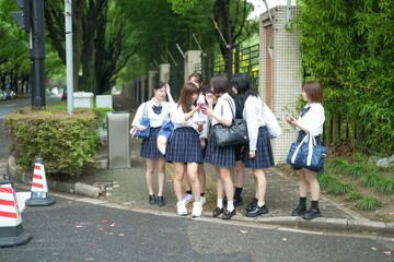 In early summer in Pudong, Shanghai, seven Japanese high school girls in summer uniforms are walking along a city street. They pause together, smiling as they look at something on a smartphone.