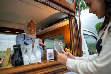Chinese muslim female barista in hijab showing qr code to indonesian southeast asian woman to scan with cellphone for payment. Mockup or copy space. Small business of coffee shop or cafe on truck