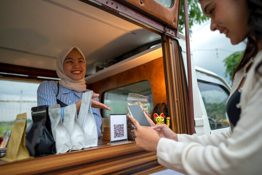Chinese muslim female barista in hijab showing qr code to indonesian southeast asian woman to scan with cellphone for payment. Mockup or copy space. Small business of coffee shop or cafe on truck