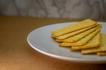 A close up of a stack of golden, rectangular crackers on a white plate, set against a brown surface and subtle textured background. Ideal for food content