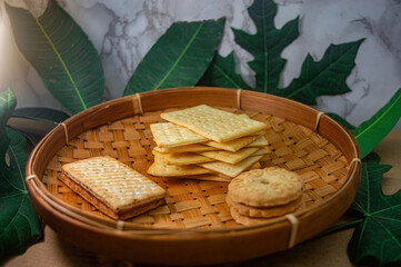 A rustic bamboo woven basket filled with various golden biscuits and crackers, accented by green leaves and a marble background. Ideal for natural snack displays