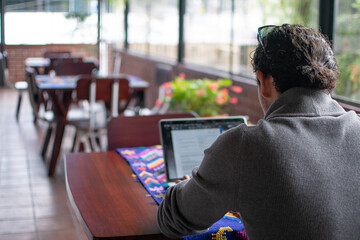 Photograph of an adult man wearing a sweater and glasses working on a laptop at a wooden table with a textile tablecloth, with other woven tables seen from behind.