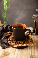 Black coffee in a black cup with coffee beans on rustic wooden table, french press on background.