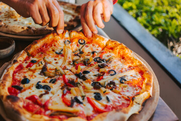 Men's hands tear off a slice of pizza with vegetable toppings, olives and stretching hot stretchy cheese. Process of eating pizza close-up. Delicious Italian pizza for friends and delivery