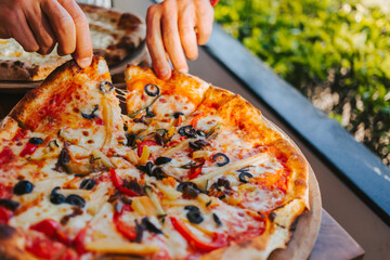 Men's hands tear off a slice of pizza with vegetable toppings, olives and stretching hot stretchy cheese. Process of eating pizza close-up. Delicious Italian pizza for friends and delivery