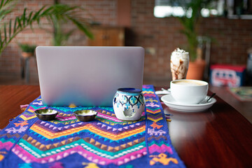Photograph of a chocolate milkshake cocktail and a large cup of coffee next to a laptop on a table with a textile-patterned tablecloth, front view.