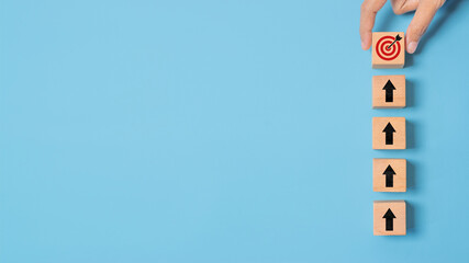 Hand holding wooden block with target aiming dart board and arrow up on blue background. Business achievement objective goal and project progress growth value processing marketing financial concept