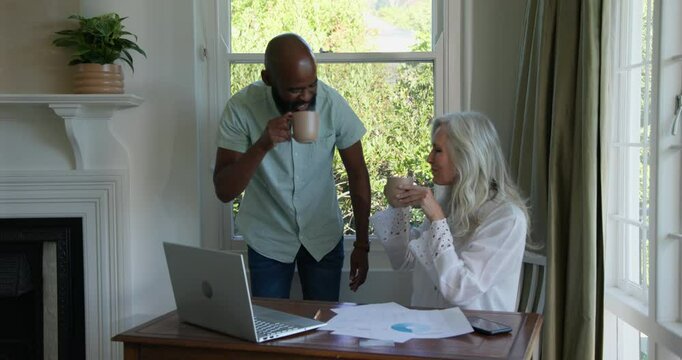 Diverse senior couple receiving feedback and celebrating win at home office with laptop and charts