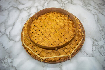 A top down view of a traditional round woven bamboo tray or tampah on a white and gray marble...