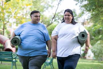 Happy Overweight indian couple holding yoga matt walking together at summer park. fat man and woman healthcare. Plus size people fitness india. Obesity concept.