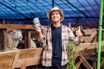 Indonesian Shepherd Showing Money and Thumb Up Against The Goat's Pen