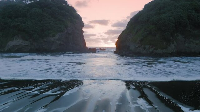 Coastal scene at sunset in Bethells Beach, New Zealand. Ocean waves crash on the black sand beach between two rocky cliffs covered in lush green vegetation. The sky is a soft pink and orange.