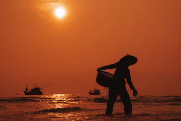 Silhouette of Vietnamese woman in straw hat on beach of sea with ships in fishing market in village in the morning at sunrise in Vietnam