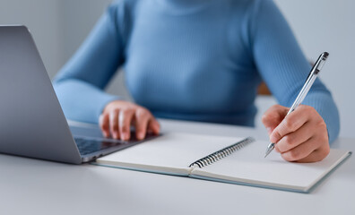 Fototapeta premium Close-up of woman taking notes and working on laptop at home office. Female student studying online, e-learning.