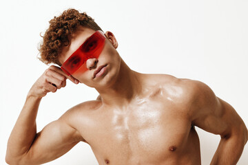 Confident shirtless young man posing with red sunglasses, showcasing a strong physique against a neutral background. Fashion and self-expression concept.
