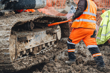 Construction worker  in bright safety gear working hard using shovel to clean tracks of his heavy machinery excavator on muddy site © Iryna