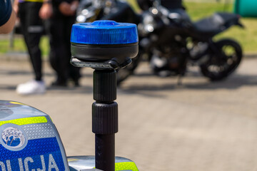 blue flashing light on a police motorcycle, with blurred people and motorcycles in the background. The word "Policja" is visible on the vehicle. © Adam