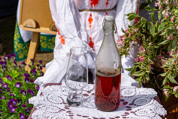 glass bottle with homemade red alcohol and a glass placed on a decorative doily. In the background, a woman in traditional folk costume and flowers create the atmosphere of a regional fair or folk eve