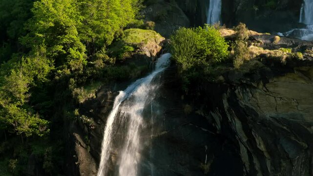 Acquafraggia waterfalls, water shimmering in sunlight as it cascades down rocks, surrounded by lush green trees, Lombardy, Italy. Aerial approach
