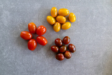 An overhead view of three groups of cherry tomatoes—yellow, red, and brown—arranged in a flower-like pattern on a gray stone surface.