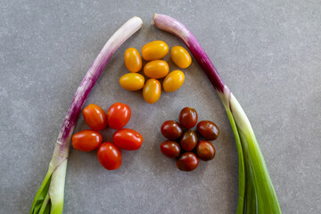 An overhead view of three groups of cherry tomatoes—yellow, red, and brown—arranged in a flower-like pattern around a bunch of green spring onions on a gray stone surface.