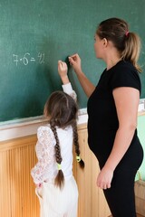 Young student works on multiplication problem on chalkboard with teacher in classroom setting,...