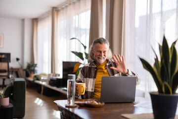 Man working from home in a modern living room, smiling and waving during a video call