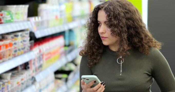 Young attractive woman checking to do list on smartphone in supermarket. Online store of hypermarkets, shopping centers. Smartphone food market girl food shopper.