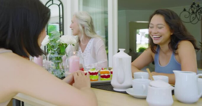 Diverse female friends initiating tea chat by teapot, senior clapping, sampling pastries on patio