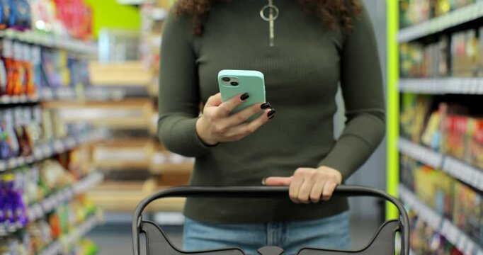 Close up of unrecognizable woman using smartphone while carrying shopping cart walking through aisles in hypermarket doing daily food shopping.