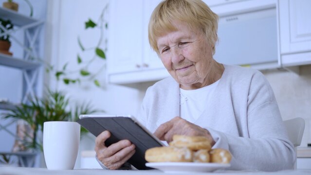 Smiling senior woman enjoying a relaxing coffee break in her modern kitchen, using a digital tablet to browse online, while savoring delicious pastries and feeling happy