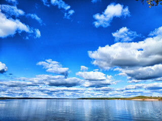 Stunning blue sky with fluffy clouds reflecting on calm lake water during summer afternoon near a scenic shoreline