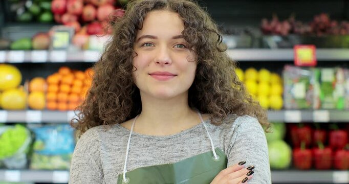 Young woman worker in a vegetable and fruits section supermarket standing smiling with arms crossed. Friendly female looking at camera in a shop market.