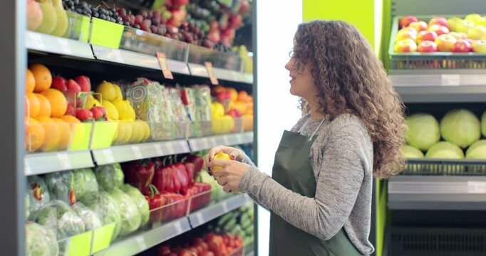 Young woman worker in green apron stocking the lemons in supermarket. Young employee at work. Curly girl arranging fruits on shelf. People and profession. Grocery shop. Consumerism.