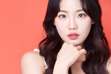 Fototapeta premium Young Asian Woman with Wavy Hair in White Sleeveless Top Touching Chin on Coral Background