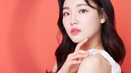 Young Asian Woman with Wavy Hair in White Sleeveless Top Touching Chin on Coral Background