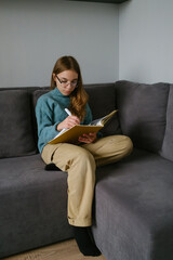 Teen girl writing in notebook on sofa, focused and relaxed in comfortable setting.