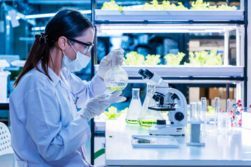 Female scientist examines green solution in a lab surrounded by plants and test tubes, representing...