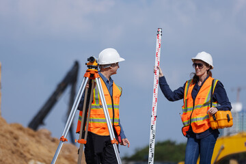 Two construction surveyors in safety gear stand at a job site using a tripod-mounted total station...