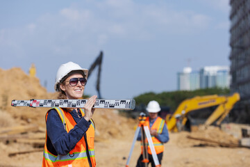 A female construction surveyor in safety gear smiles while holding a leveling rod on her shoulder...