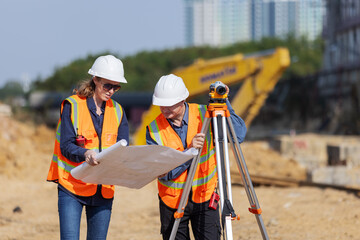 Two construction surveyors in safety gear work at a job site, with one reviewing blueprints and the...
