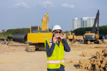 A female construction engineer in a hard hat and reflective vest adjusts her headset at a busy job...