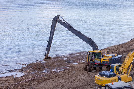 Long-arm excavator dredging riverbank with construction equipment nearby.