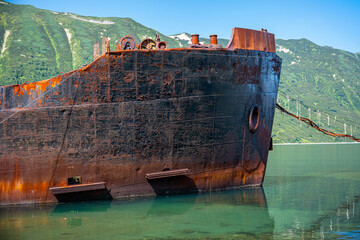Rusted shipwrecks in clear blue water with rocky shoreline and green hills in background under...